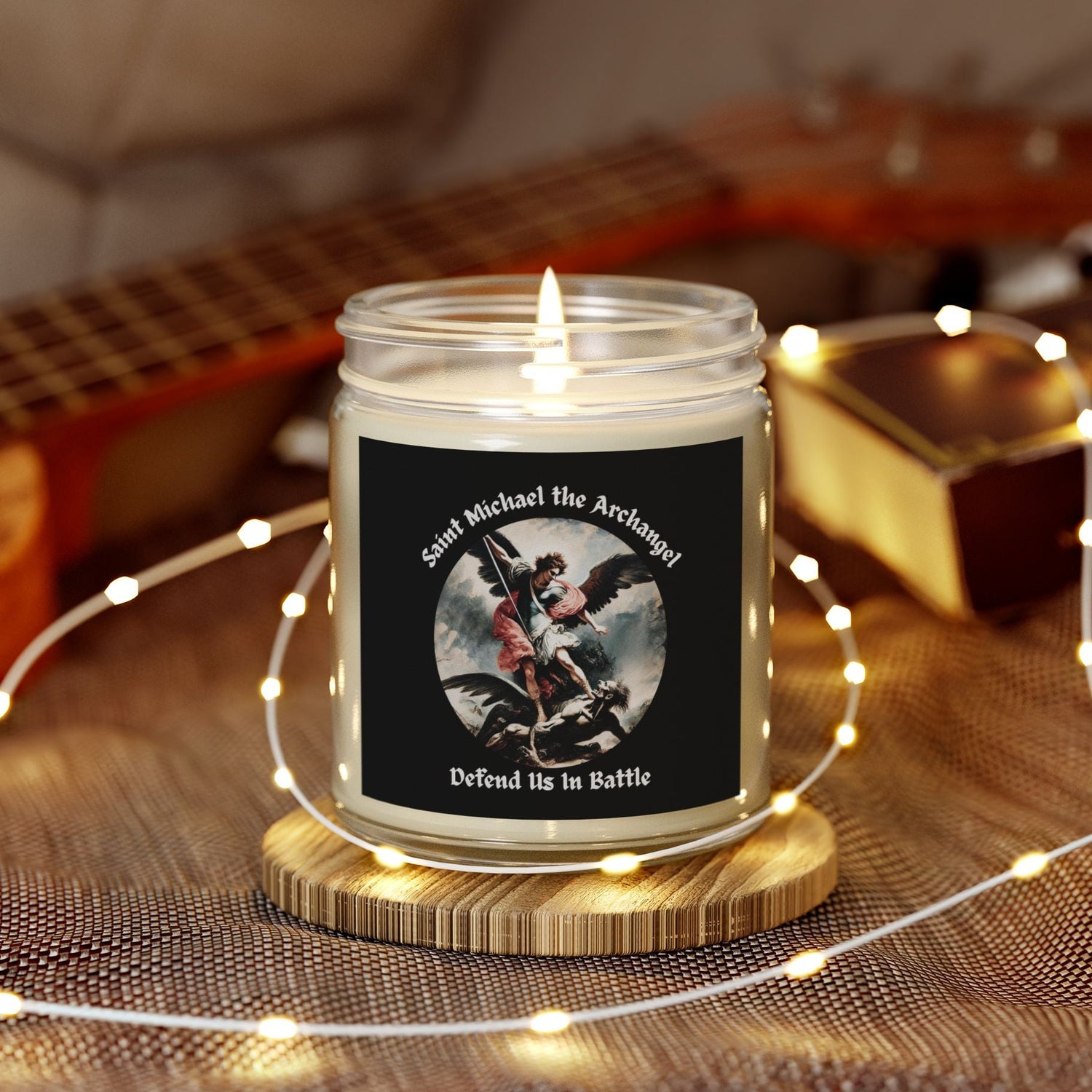 Saint Michael Design Candle in a glass jar with a decorative label on a wooden coaster, surrounded by string lights and a guitar in the background.